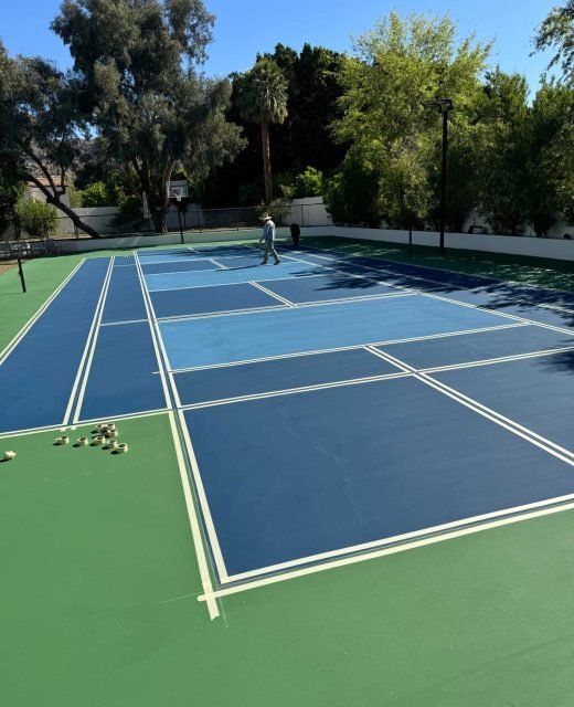 Bright blue tennis court with freshly painted white lines, surrounded by lush greenery, under clear sky.
