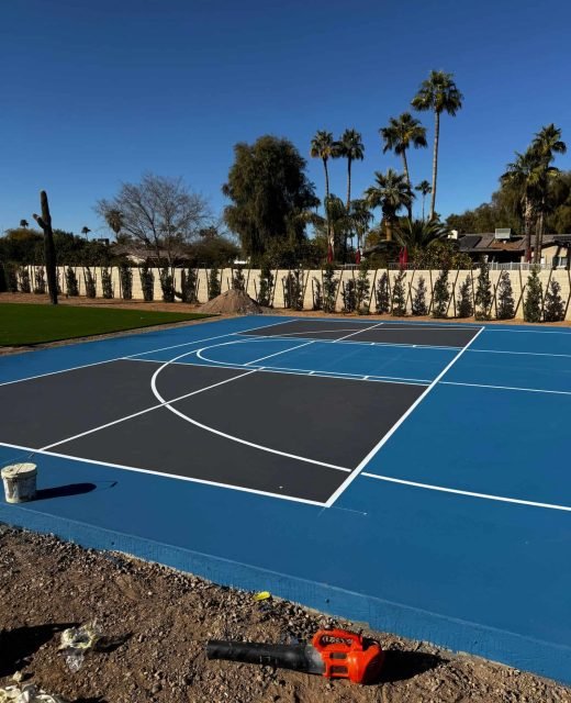 High-quality image of a newly painted basketball court with blue and black surface, white boundary lines, under clear sky for sports facility improvement.